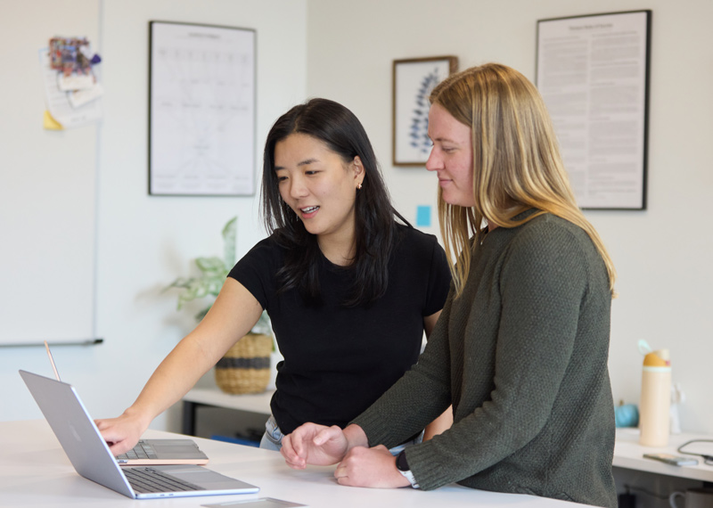 Two students talk to each other, pointing to something on a laptop.
